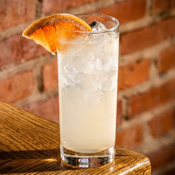 Tall glass cocktail with ice and a grapefruit slice on a wooden bar against a brick wall.