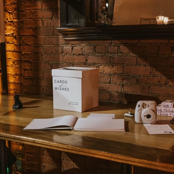 A "Cards and Wishes" box, guestbook, and instant camera on a wooden table against a brick wall.