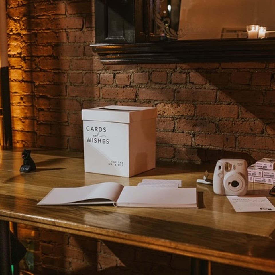 A "Cards and Wishes" box, guestbook, and instant camera on a wooden table against a brick wall.