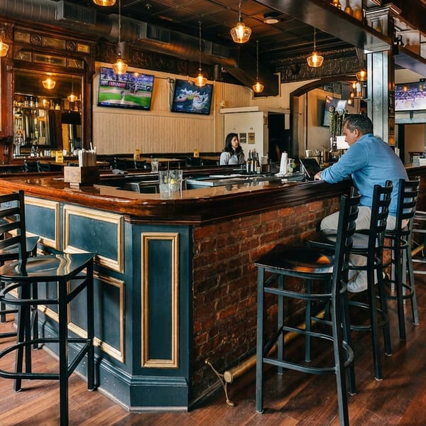 Cozy bar interior with dark wood, brick, ornate mirrors, bar stools, and patrons watching sports on TVs.