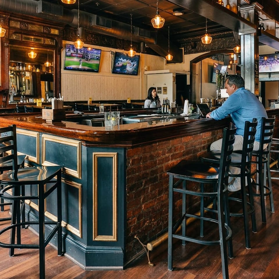 Warmly lit restaurant bar with a man seated, ornate wood paneling, and multiple televisions.