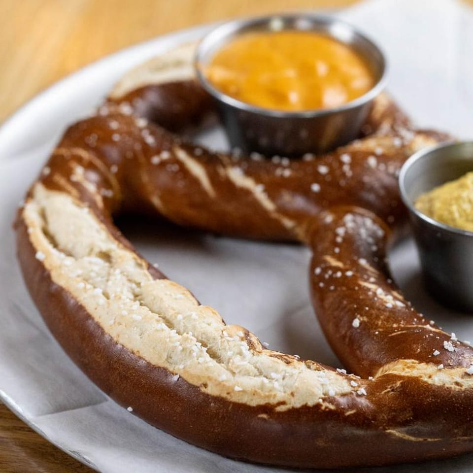 A large, golden-brown salted pretzel with cheese dip and mustard on a white liner.