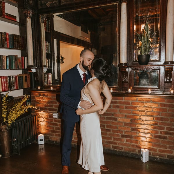A couple kisses in a dimly lit room with a large mirror, brick wall, and bookshelves.