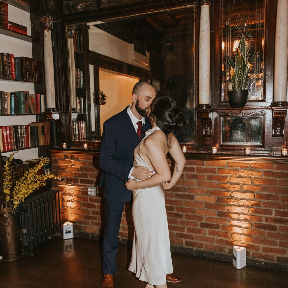 A couple kisses in a dimly lit room with a large mirror, brick wall, and bookshelves.