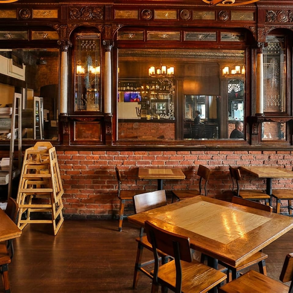 Restaurant interior with wooden tables, chairs, brick wall, ornate wood, and mirrors reflecting a bar.