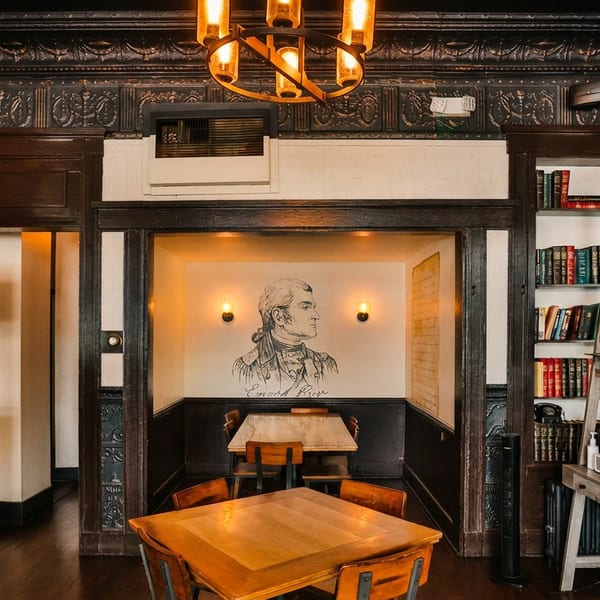 Warmly lit restaurant interior with dark wood, brick, tables, bookshelves, and a historical figure mural.
