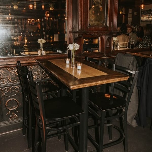 A cozy, dimly lit bar interior with a high-top table, black chairs, and a large mirror reflecting warm lights.