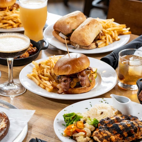 A table laden with burgers, fries, chicken wings, grilled chicken, pretzel, and various drinks.
