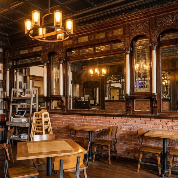 Cozy restaurant interior with wooden tables, exposed brick, ornate mirrors, and bookshelves under warm chandeliers.