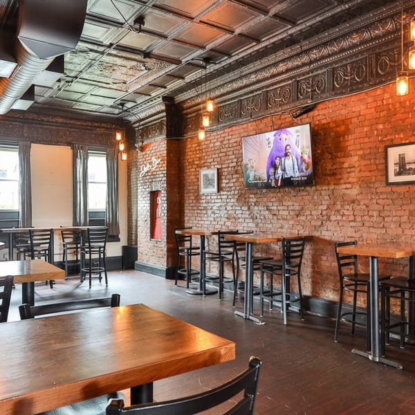Rustic restaurant interior featuring brick walls, wooden tables, black chairs, and a decorative tin ceiling.